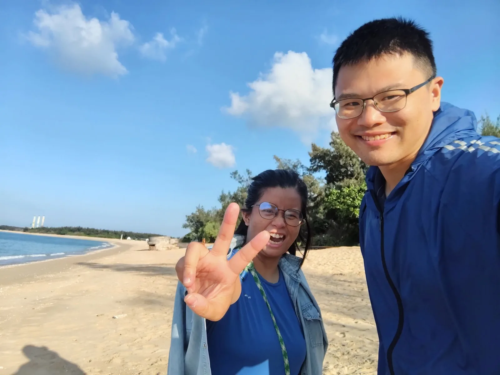 心安與琬珊在海邊的自拍合照，左側是藍天、海岸與沙灘，琬珊比著勝利手勢