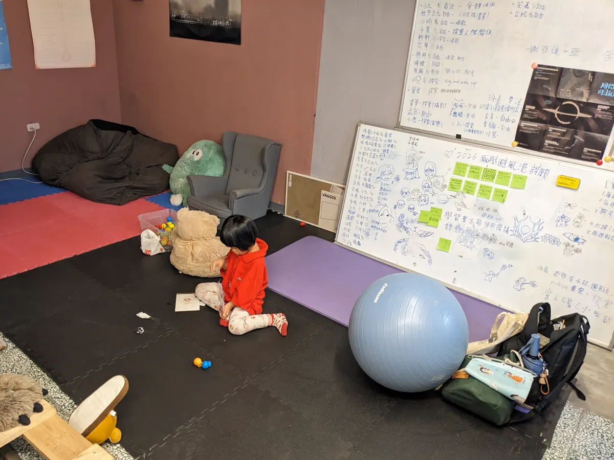 避風港的主要空間：軟墊地板、懶骨頭、布偶與寫滿字的白板，一個孩子坐在地上玩